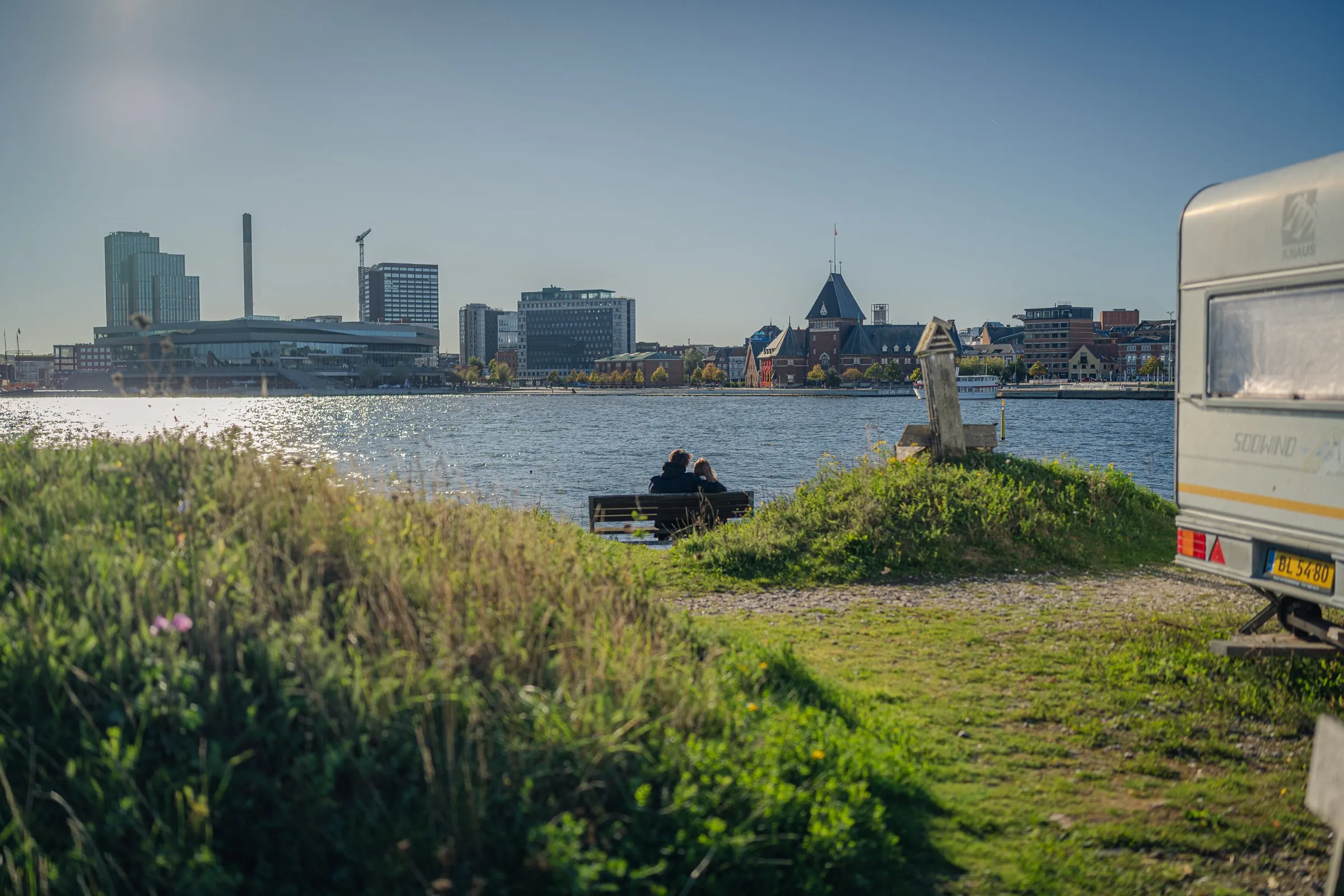 Grassy mounds by the water, a caravan, the city skyline opposite