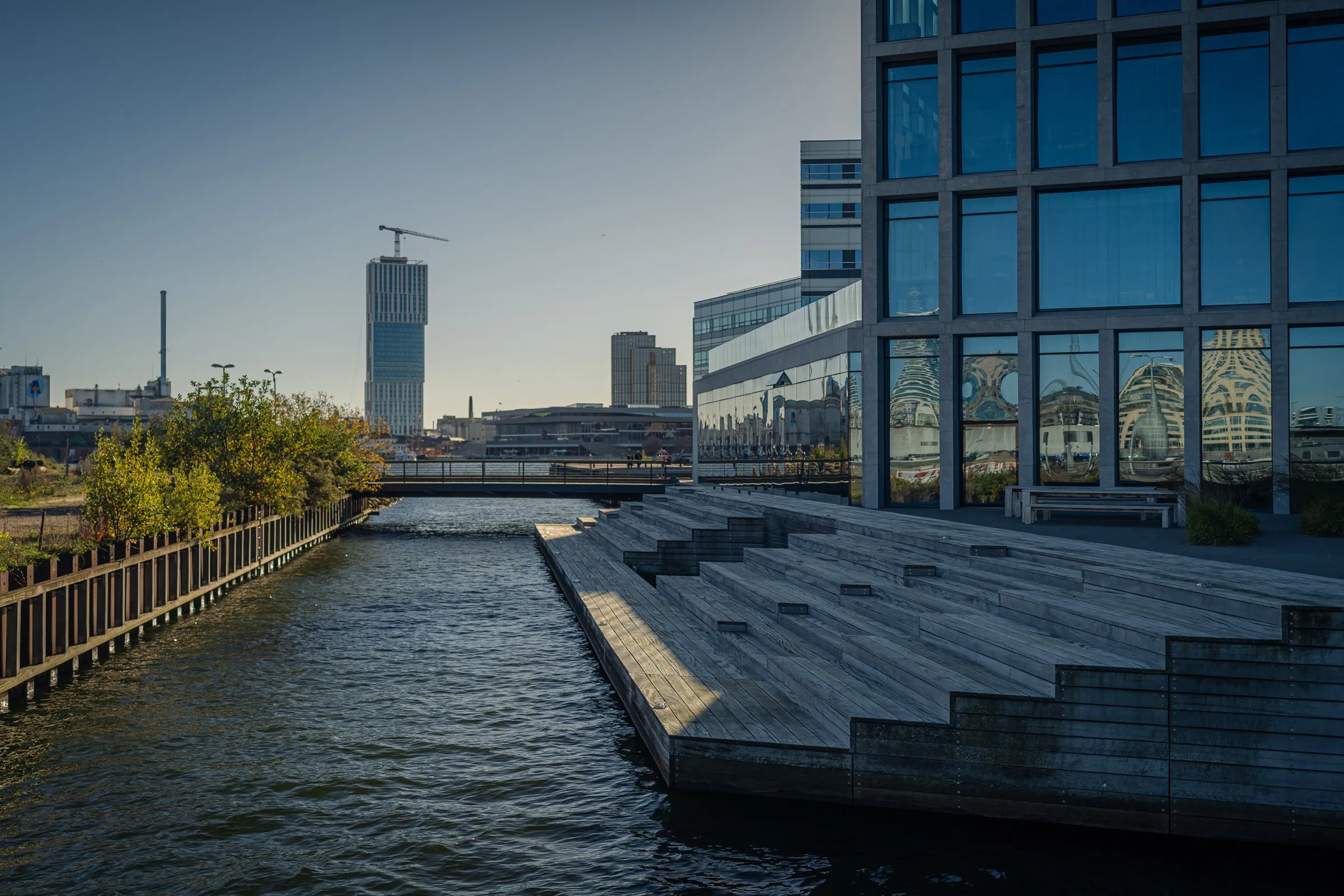 A canal with timber seating steps, glass buildings on the right, the Lighthouse tower and a crane behind