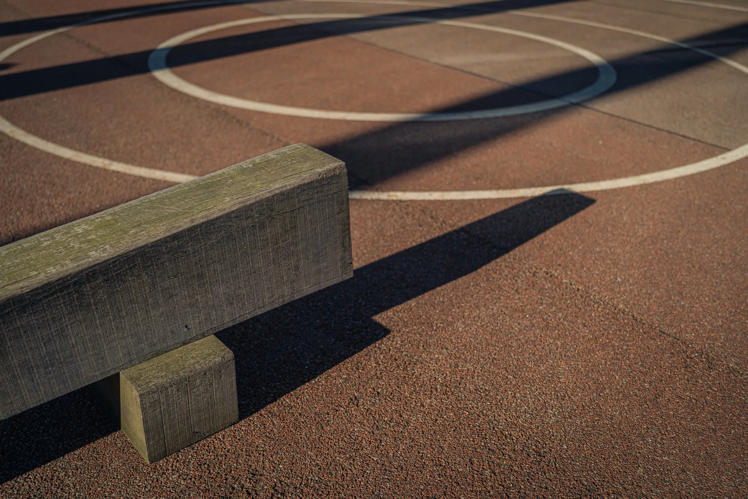 A timber beam on red sports surfacing with white lines, a long shadow