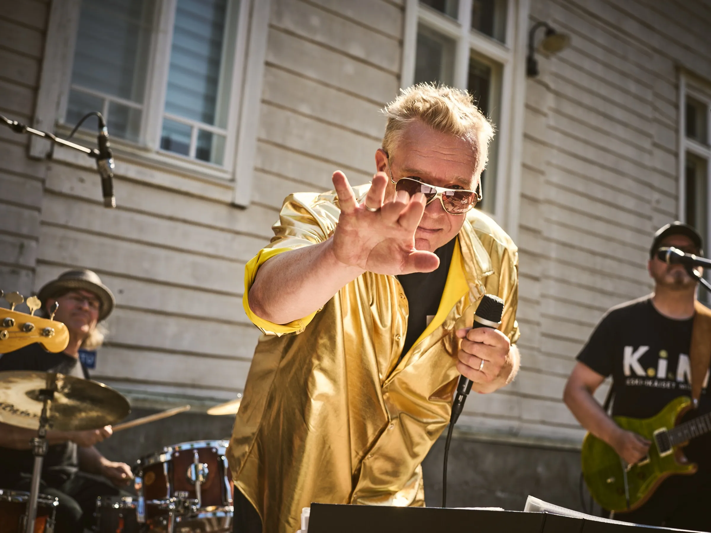 Singer in a golden shirt reaching a hand toward the camera, band in backlight