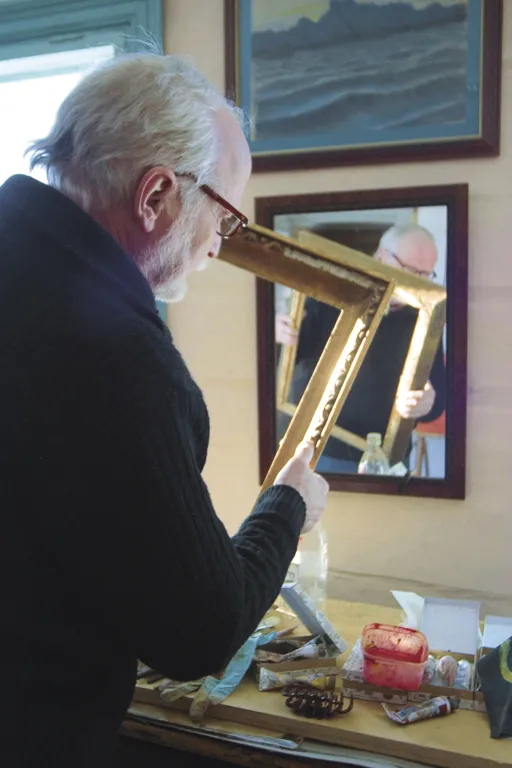 Seppo Tamminen examining a golden picture frame in his workshop, his face appearing in the mirror behind