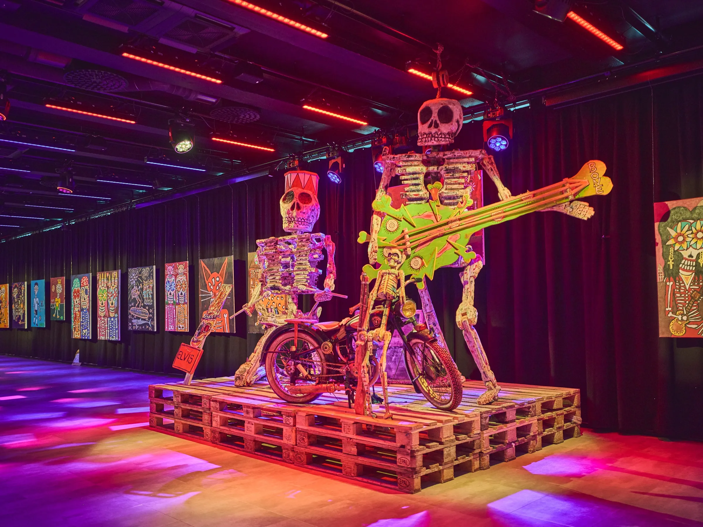 Skeleton figure with a yellow guitar next to a skeleton on a bike, painted wood