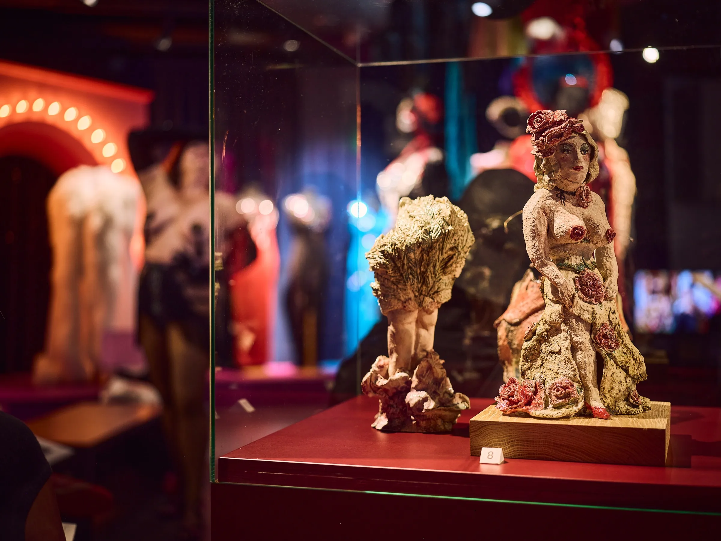 Two small ceramic figures in a display case, behind them the colorfully lit stage of the exhibition