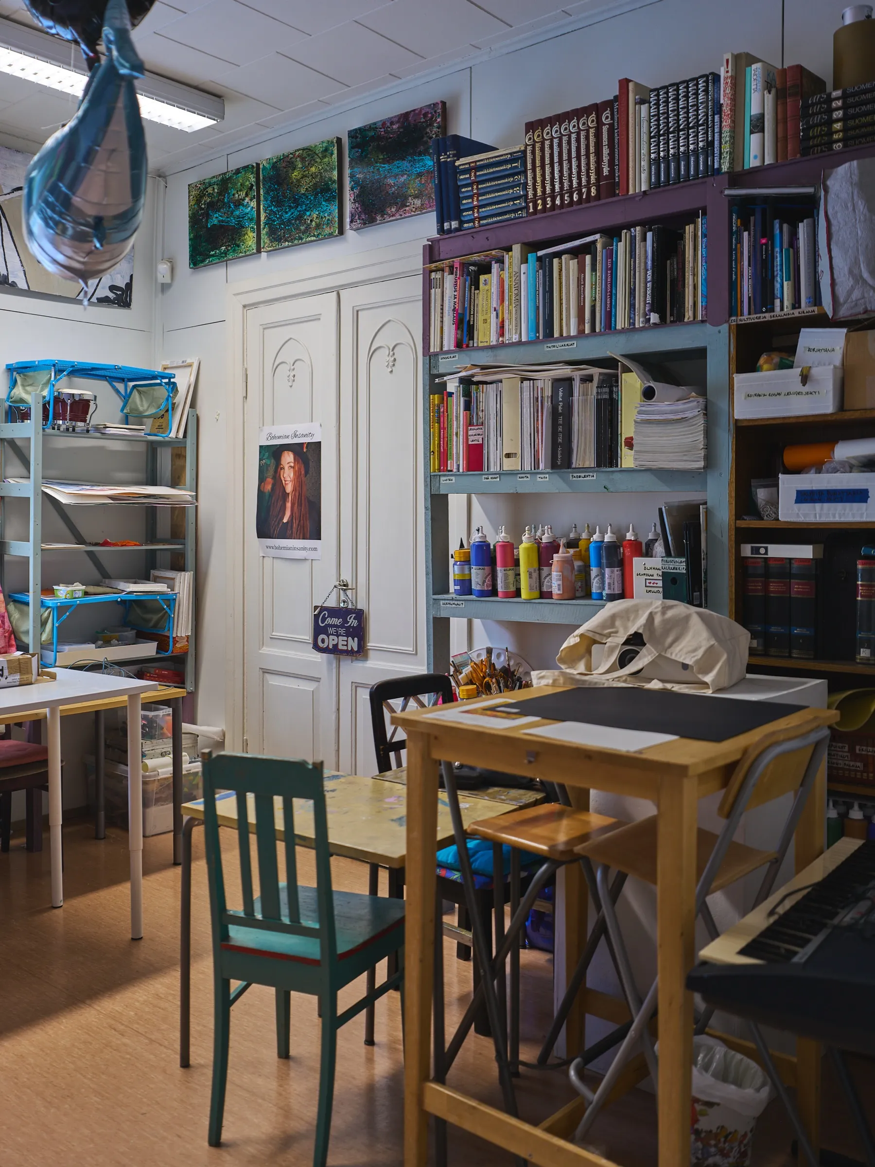 Workroom with paint bottles, bookshelves, a keyboard and work tables under three small paintings